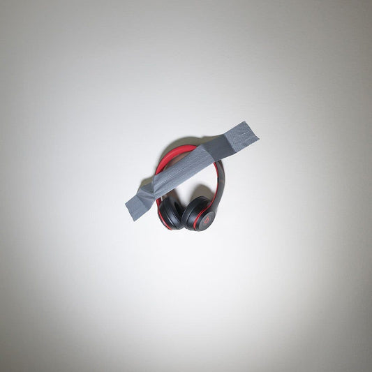 The Mediocre Headphones on Wall by headphones.com features black and red headphones taped to a white wall with gray duct tape across the headband, creating a quirky art piece.