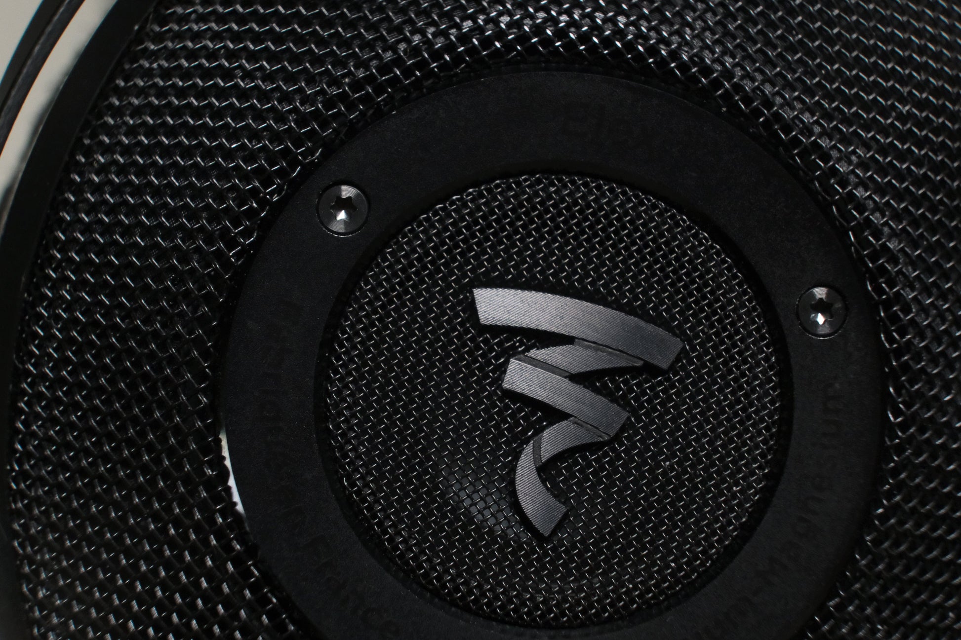 Close-up of a textured black speaker grille with a metallic Focal logo at the center, echoing the abstract design of Focal Elex Headphones. Two screws on the black ring frame the logo, underlining Focals dedication to sonic perfection.