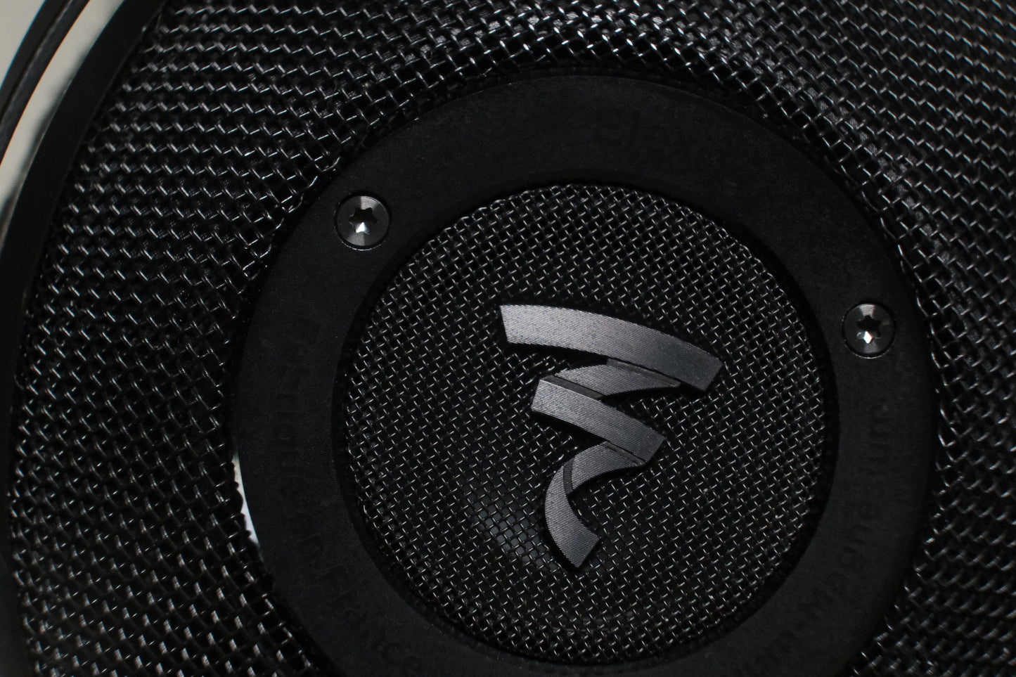 Close-up of a textured black speaker grille with a metallic Focal logo at the center, echoing the abstract design of Focal Elex Headphones. Two screws on the black ring frame the logo, underlining Focals dedication to sonic perfection.
