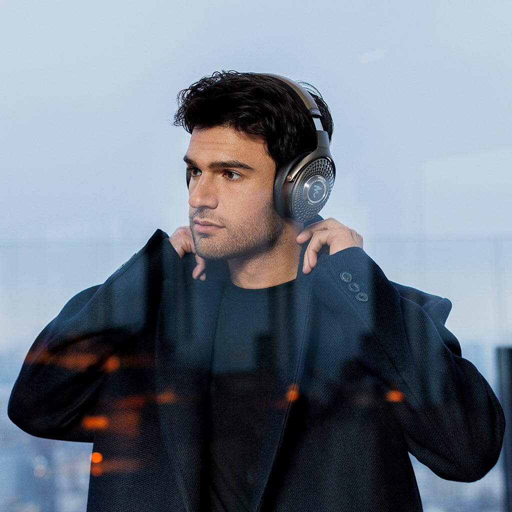 A man in a dark coat stands indoors, adjusting his Focal Bathys MG Wireless Headphones. Through the window behind him, a cityscape appears amid subtle reflections.