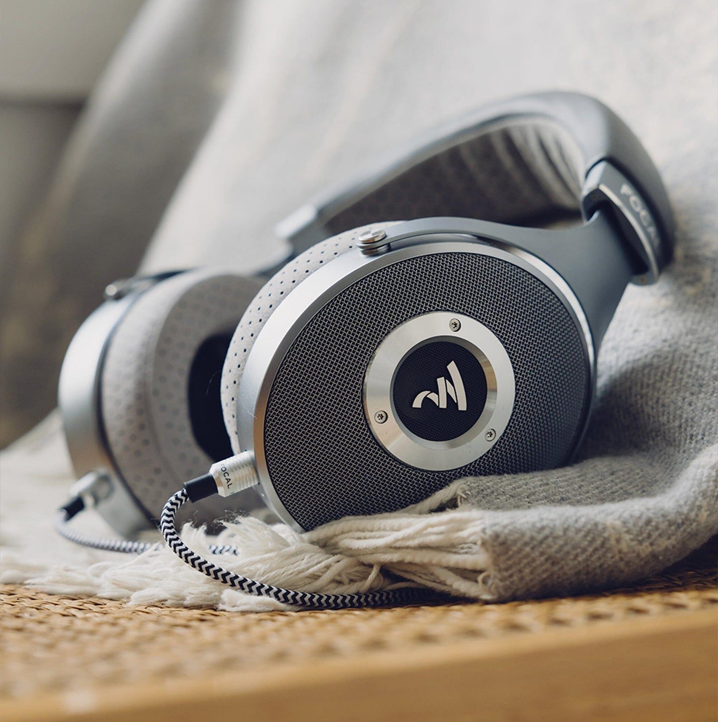 A close-up of Focal Clear Headphones by Focal, featuring a circular metal design and braided cable, resting on textured gray fabric and wicker—available now at headphones.com.