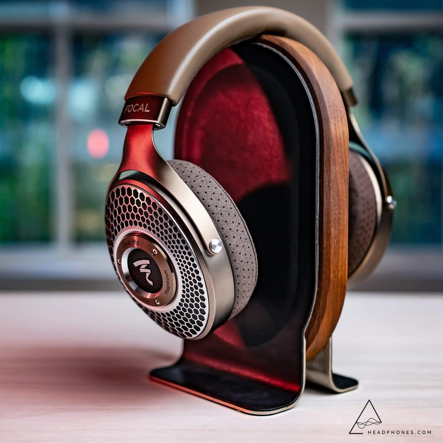 A pair of Focal Clear Mg Headphones by Focal with open-back, perforated ear cups sit on a wooden and metal stand atop a light surface, with a blurred window in the background.