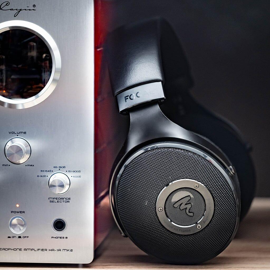 A pair of Focal Elex Headphones by Focal rests against a silver tube headphone amplifier with glowing tubes and control knobs, offering a glimpse of sonic perfection against a dark, blurred background.
