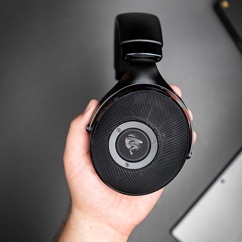 A hand holds the Focal Elex Headphones by Focal, showcasing their mesh earcup design and circular logo, set against a dark gray backdrop.
